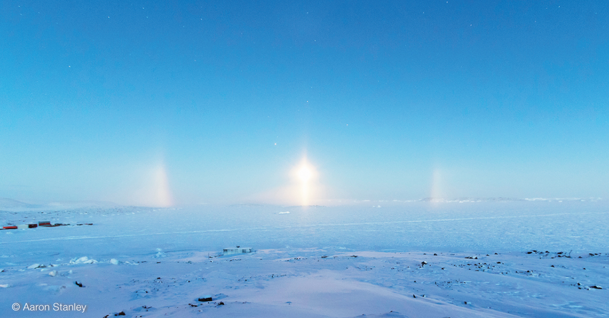 Gleaming light pillar with the lower edges of a halo to either side, over the ice, which appears blue as it's reflecting the blue sky.