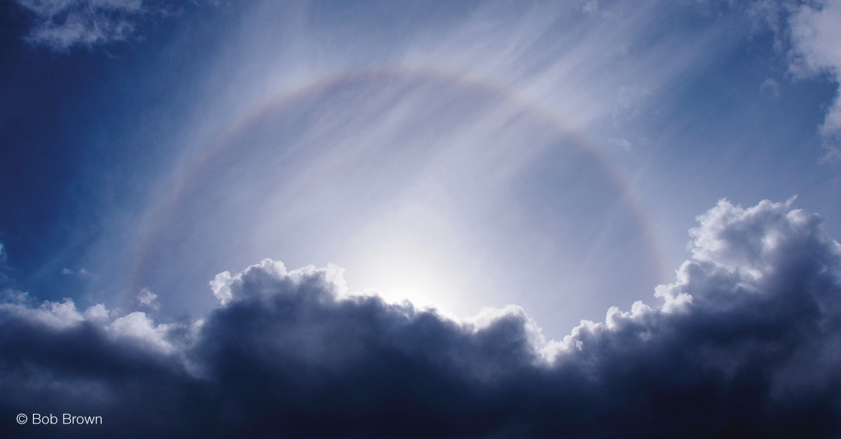 Semi-circular halo rising from dark grey clouds in a blue sky.