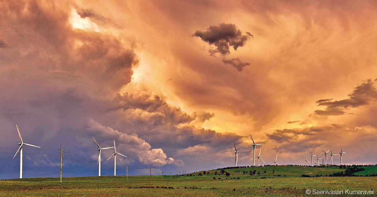 Varied smooth and rough grey clouds over wind turbines on a green hillside.