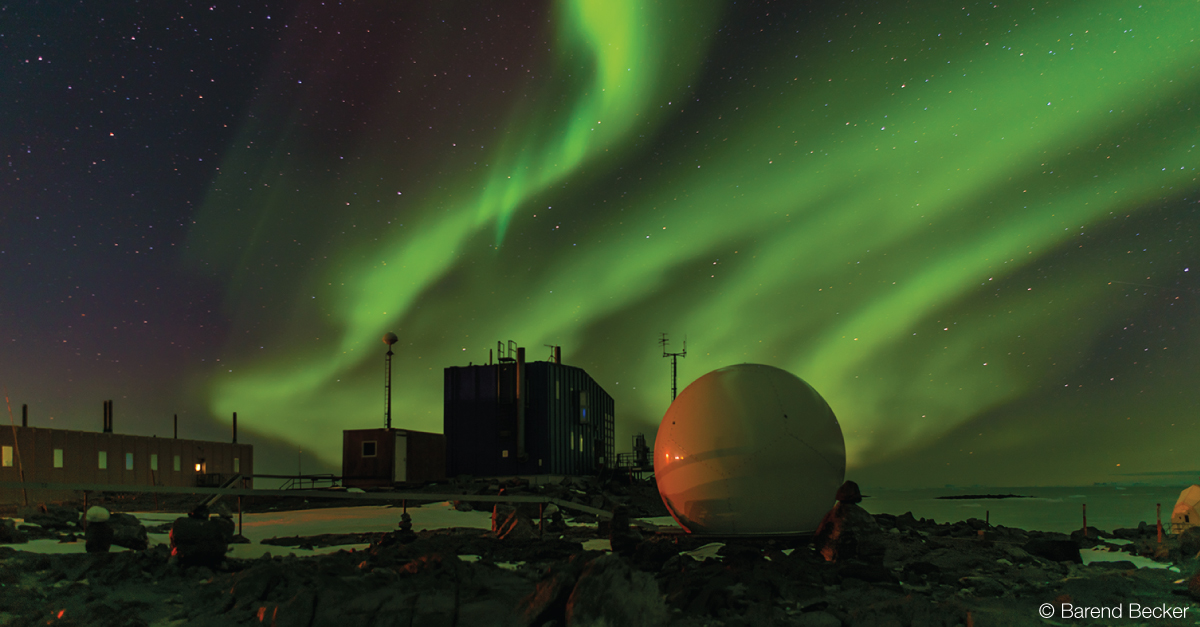 Bright green waves of aurora in the night sky, with stars dotting around and in it and a round object in the foreground, with building silhouetted in the background