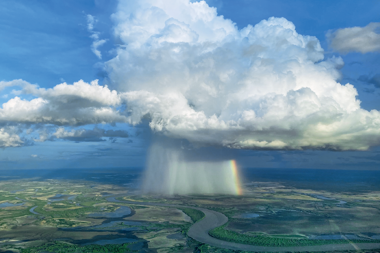 Aerial view of a tall white cloud with a column of heavy rain falling on the land below and clear skies around it. There's a rainbow in the rain.