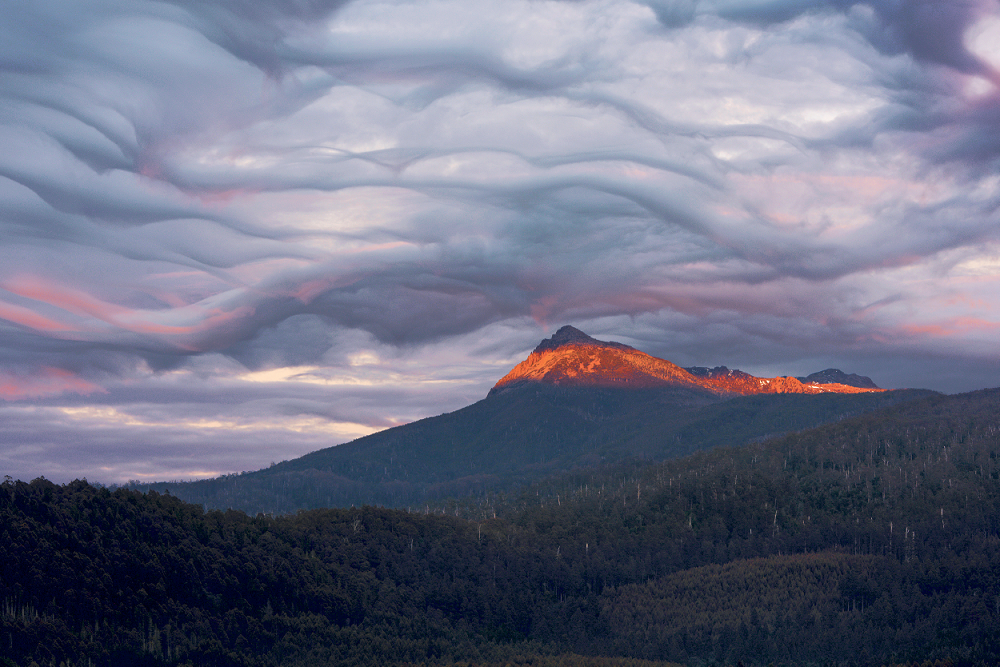 Wavy grey cloud above a mountain. The sun tinges the mountain top and sky with pink.