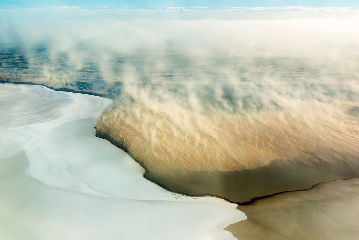 A wispy white cloud of salt dust hovers above a white lake and sandy ground.