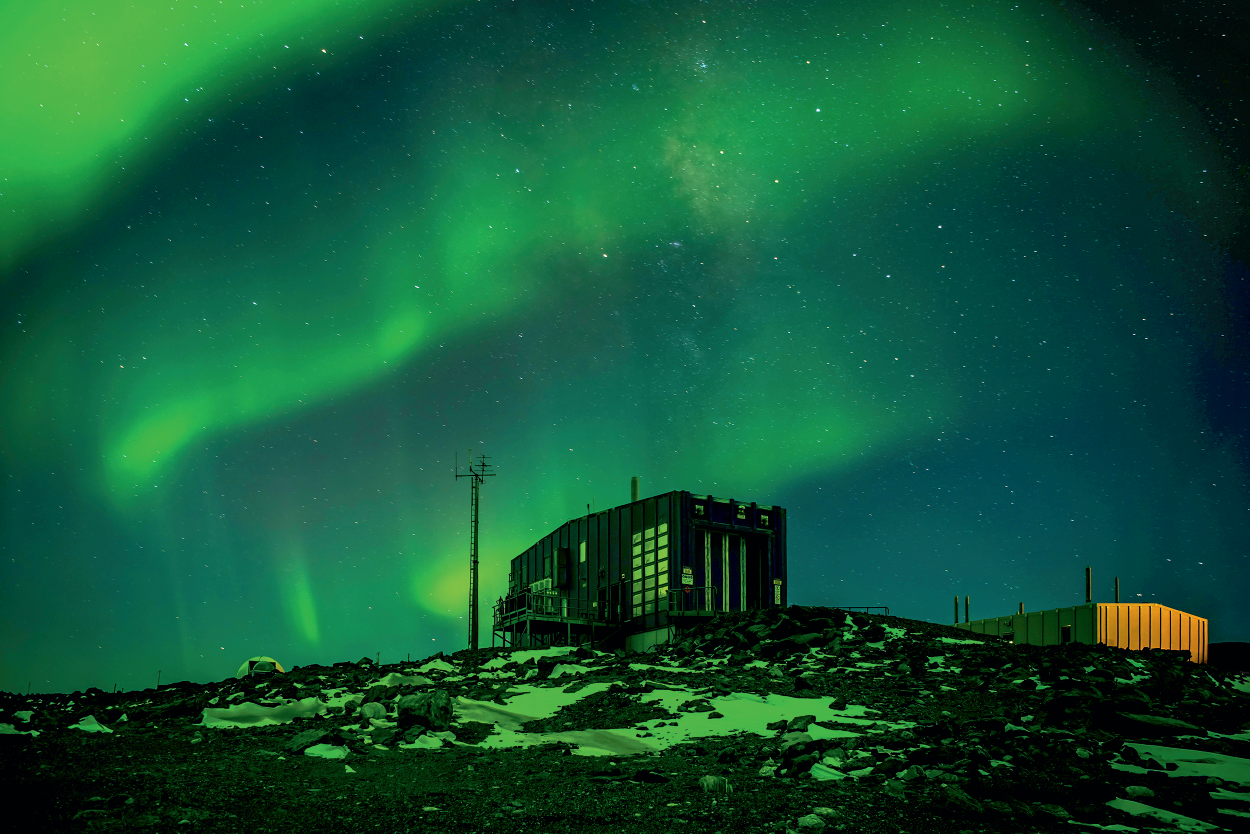Waves of green light in the night sky above a building. Green is also reflected in the snowy ground.