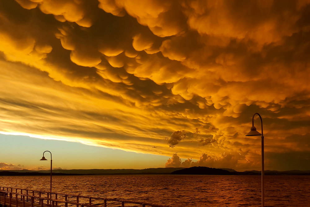 Bobbly grey mammatus cloud hangs down from a yellow sunset sky over the sea, with street lamps in the foreground.