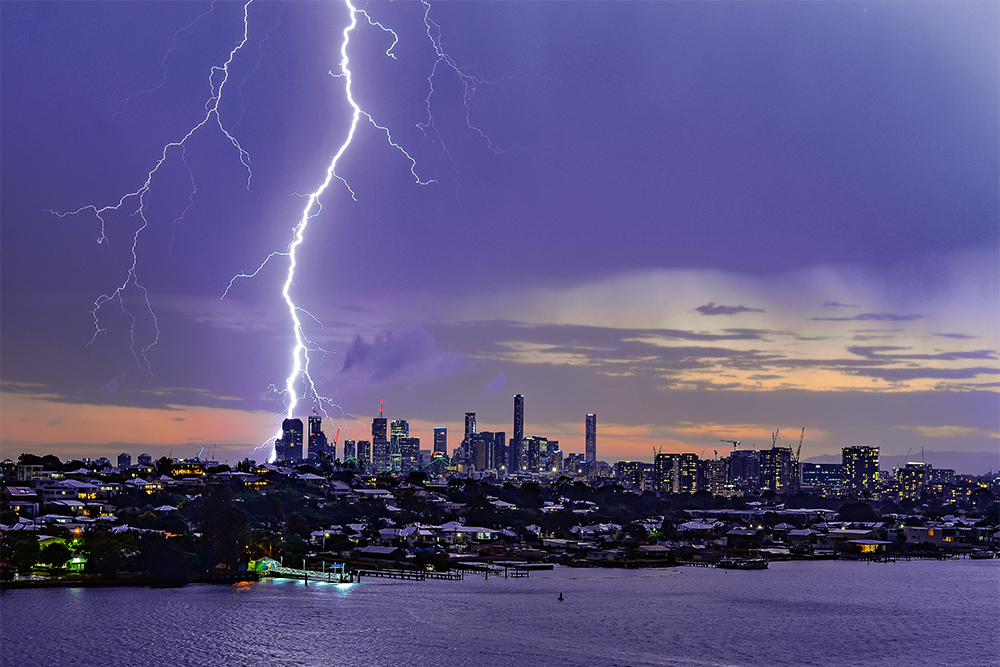 A bright shaft of lightning snakes down a dark sky over the skyline of Brisbane CBD. Secondary bolts (less bright) are to the right and left of the major one.