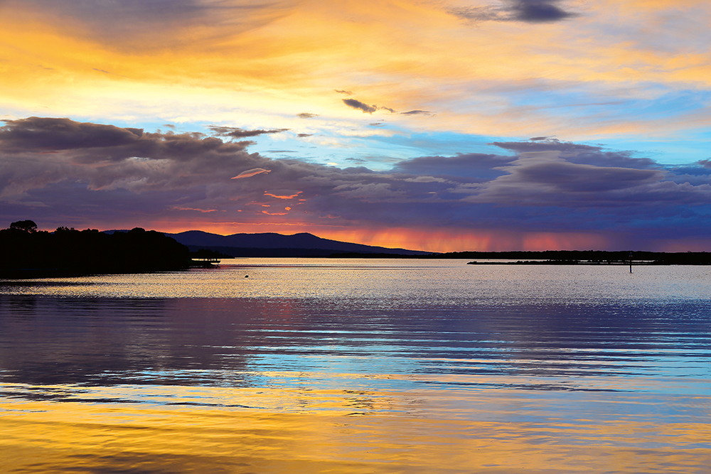 Colourful sunrise sky with grey clouds, gold, blue, orange and red is reflected in the still sea, with low hills in silhouette at the shore.