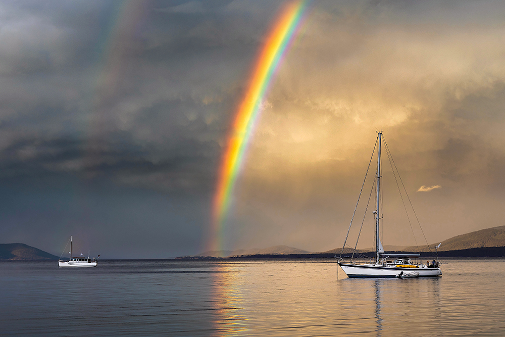 Double rainbow in grey cloudy sky over single-masted boat in the sea. The brighter rainbow is reflecting in the water.