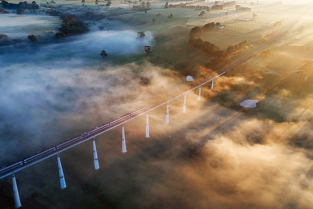 An aerial image of a fog-filled valley, with the sun casting long rays through it and a train crossing a bridge.
