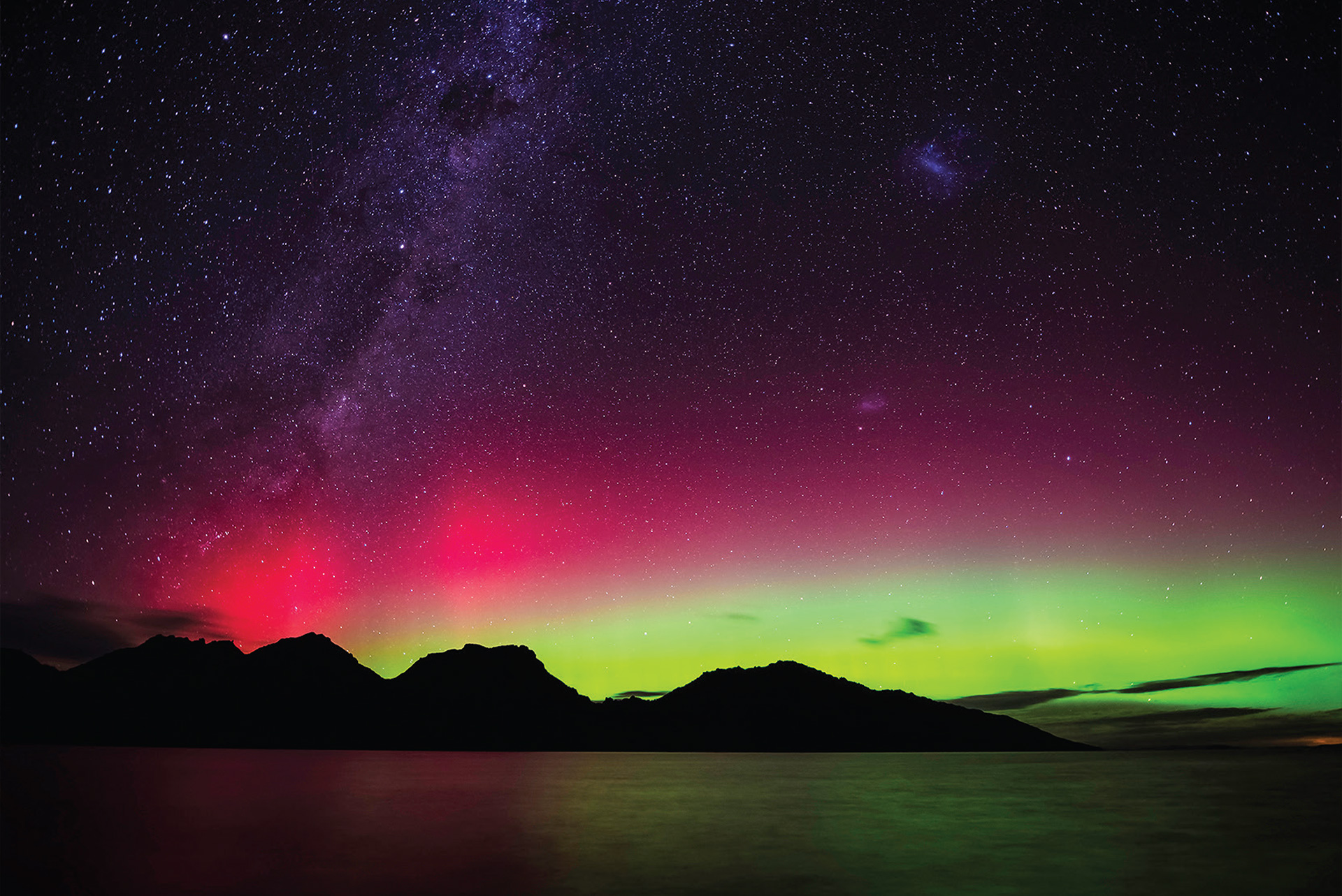 Purple, pink and green layers of light in the night sky, over silhouetted hills and sea, which reflects the colours