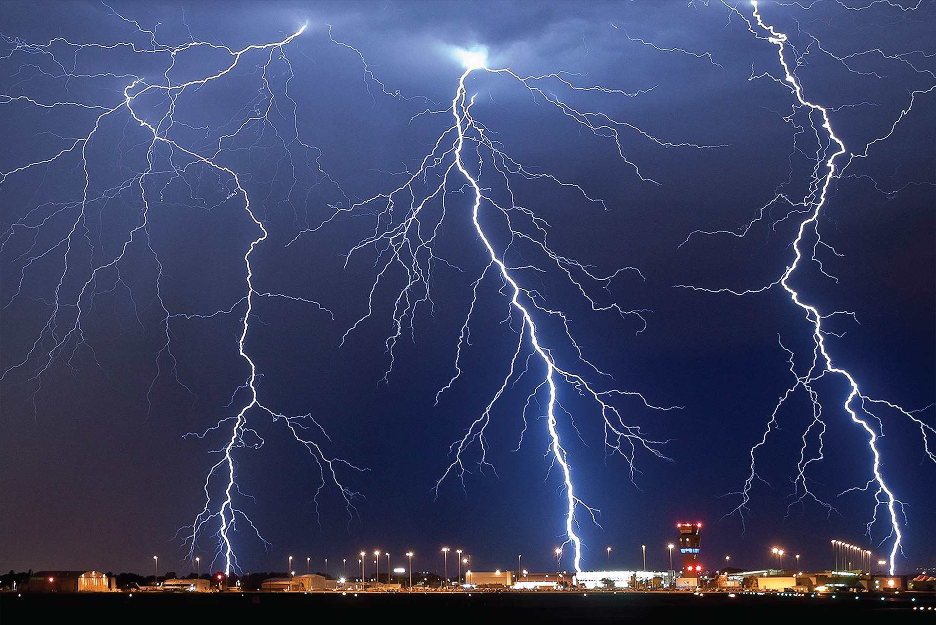 Three branching lightning strikes in a night sky over airport building and control tower