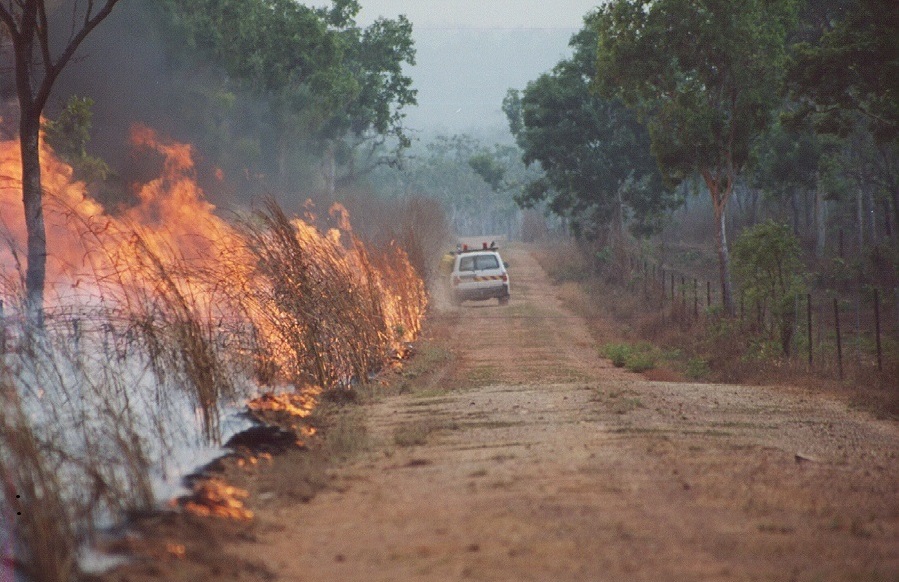 Dirt road with fire burning in low brush and trees on the left side of the road. Four-wheel drive vehicle with people in high vis down the road.