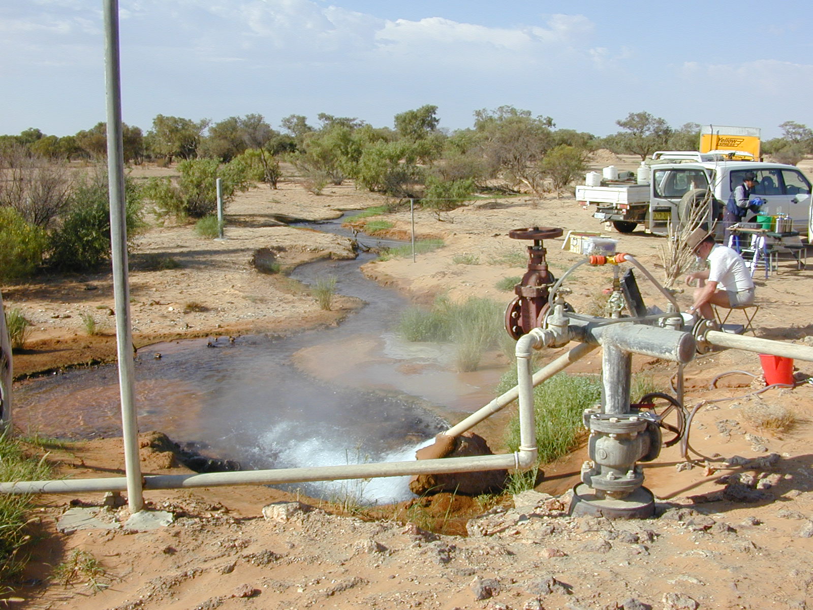 Machine with pipe in the waters of a creek in dry landscape with trees. A man tends to equipment nearby.