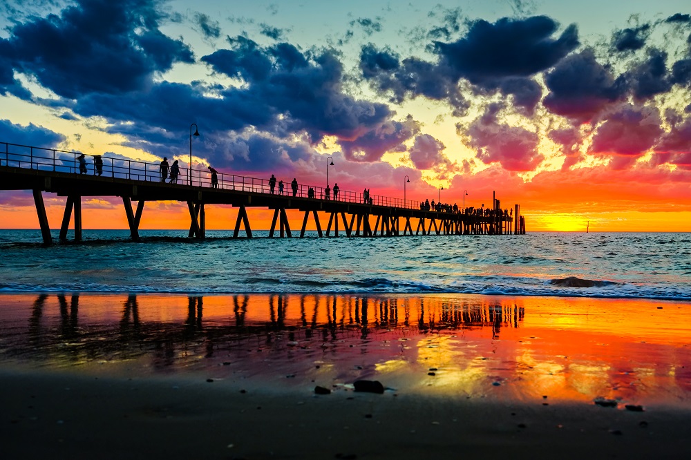 Sunset photo of people walking on a pier with the orange and gold colours of the sky reflected in the shallows