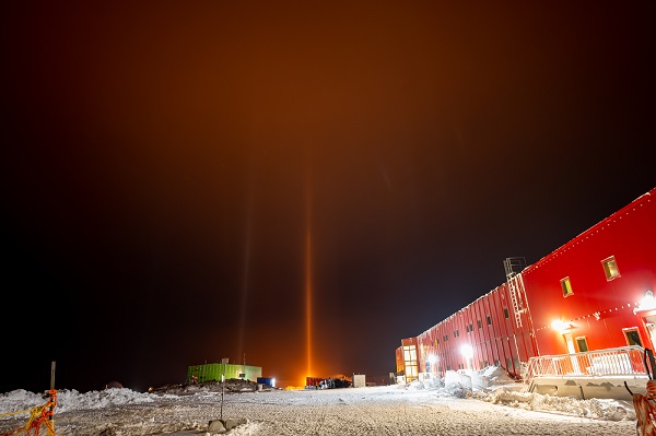 Orange light pillars reach down from the night sky to the snowy ground and buildings at Casey station, Antarctica.