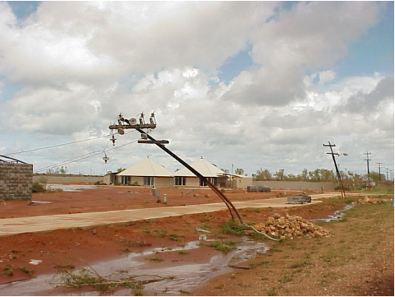 A steel power pole is bent down over the footpath by the force of the wind. The sky is cloudy and the earth red; there's a couple of houses in the background.
