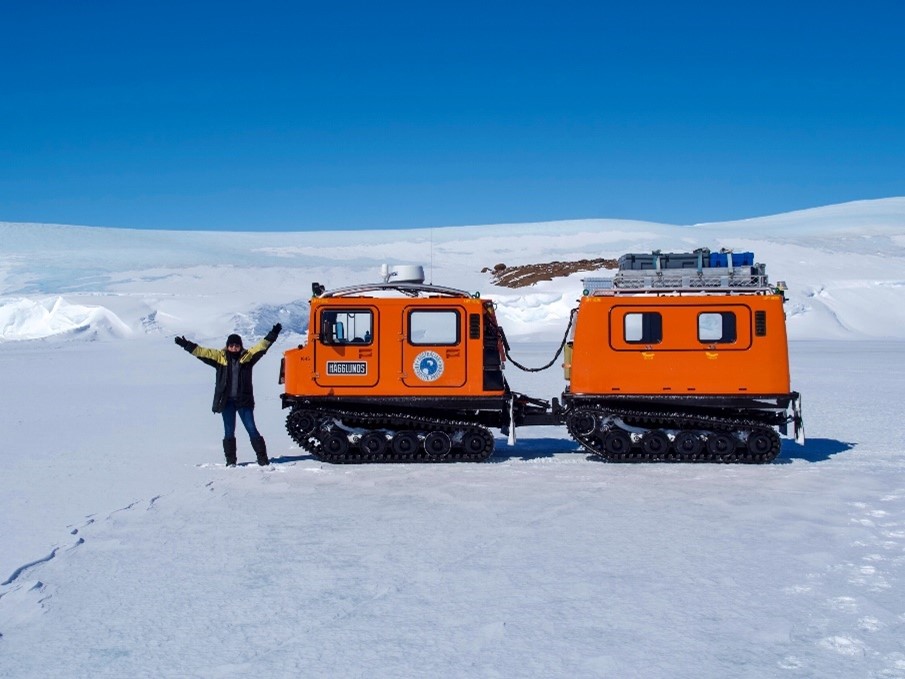 One of our technical officers,Alana-Jayne, standing in front of a bright orange Antarctic vehicle with her arms raised after driving across sea ice at Mawson station.