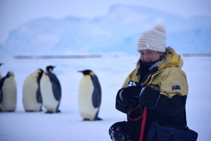 A technical officer, Alana-Jayne, sits in front of a group of emperor penguins, with ice rising upwards in the background.