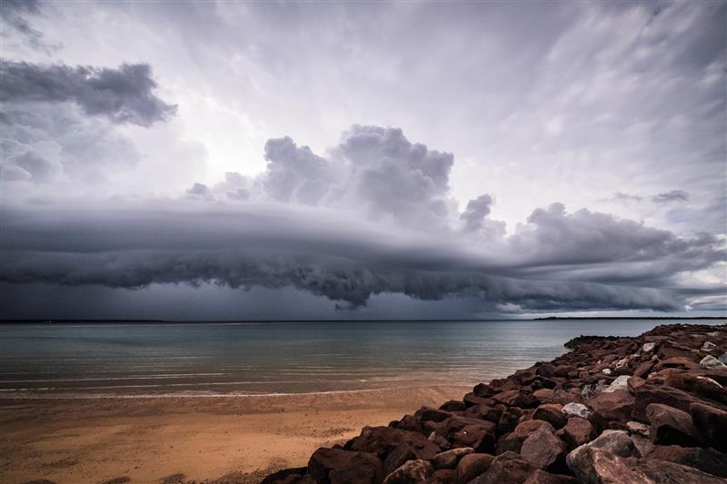 Storm clouds in a grey sky over a flat blue sea with sand and rocky shore in foreground.